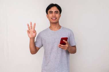 Akıllı telefon tutan gülümseyen Asyalı genç bir adamın portresi ve beyaz izole edilmiş arka planda üç parmak hareketi gösteriyor. Teknoloji, iletişim ve kampanya reklamları için mükemmel.