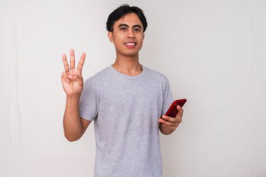 Akıllı telefon tutan gülümseyen Asyalı genç bir adamın portresi ve beyaz izole edilmiş arka planda üç parmak hareketi gösteriyor. Teknoloji, iletişim ve kampanya reklamları için mükemmel.