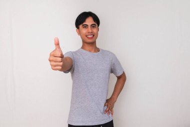 Genç Asyalı bir adamın portresi gülümsüyor ve beyaz bir arka planda izole edilmiş dostça bir şekilde baş parmak işareti gösteriyor. Reklam ve tanıtım için mükemmel.