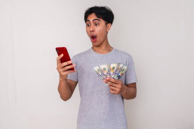 Elinde para tutarken akıllı telefona bakan, beyaz arka planda izole edilmiş, komik, genç Asyalı bir adam. Çevrimiçi alışveriş veya finansal uygulama reklamı için mükemmel