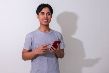 Beyaz arka planda akıllı telefonunu kullanırken gülümseyen genç Güneydoğu Asyalı bir adamın portresi dijital reklamlar, promosyonlar veya modern iletişim temaları için mükemmel.