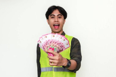 Asyalı mutlu bir inşaat işçisinin portresi, beyaz arka planda izole edilmiş, neşeli bir gülümsemeyle nakit tutuyor. Maaş, ödeme veya iş başarısı reklamları için mükemmel.