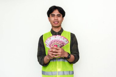 Asyalı mutlu bir inşaat işçisinin portresi, beyaz arka planda izole edilmiş, neşeli bir gülümsemeyle nakit tutuyor. Maaş, ödeme veya iş başarısı reklamları için mükemmel.