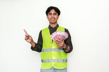 Asyalı mutlu inşaat işçisi elinde para tutarken gülümsüyor ve yanlamasına işaret ediyor, beyaz arka planda soyutlanmış. Maaş, başarı ve reklam temaları için mükemmel