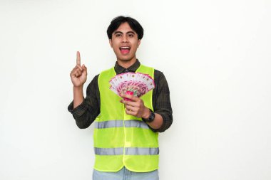 Asyalı mutlu inşaat işçisi elinde para tutarken gülümsüyor ve yukarıya bakıyor, beyaz arka planda soyutlanmış. Başarı, gelir, ikramiye veya mali başarı konuları için harika