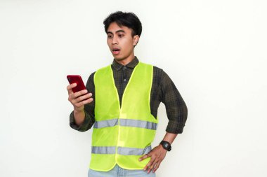 Güvenlik yeleği giymiş Asyalı inşaat işçisi akıllı telefonunu karışık bir ifadeyle tutuyor. İzole edilmiş portre bir iş, teknoloji ve endüstri anını yakalıyor.