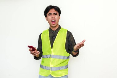 Güvenlik yeleği giymiş Asyalı inşaat işçisi elinde kızgın bir ifadeyle akıllı bir telefon tutuyor. Sektörde hayal kırıklığına uğramış bir anı gösteren izole bir portre.