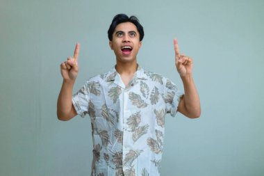 Yaz kıyafetleri içinde mutlu, genç Asyalı bir adam gülümseyip arka planda iki eliyle işaret ediyor. Heyecan ve pozitiflik gösteriyor. Terfi ya da reklam için mükemmel.
