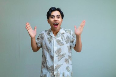 Yazlık giysiler içinde genç bir Asyalı adam portresi. Şaşırmış bir el hareketiyle, açık bir arkaplan karşısında poz veriyor. Terfi veya reklam için mükemmel.