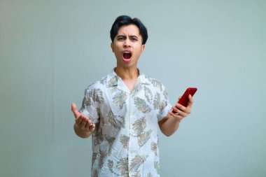 Yazlık kıyafetleri içinde, elinde akıllı bir telefonla kameraya ciddi bir ifadeyle bakan, temiz bir stüdyoda izole edilmiş kızgın, genç bir Asyalı.