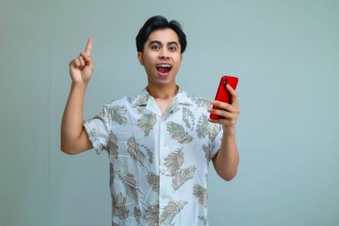 Heyecanlı, yaz kıyafetleri içinde genç bir Asyalı elinde cep telefonu, temiz bir arkaplan, neşeli bir ruh hali ve neşeli bir tatil enerjisi ile ayakta duruyor.