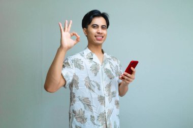 Yazlık giysiler içinde gülümseyen Asyalı genç bir adamın portresi. Akıllı telefon tutarken ve kullanırken el işareti yapıyor. Neşeli bir arkaplan karşısında duruyor.