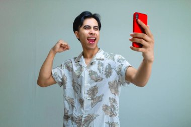 Yazlık kıyafetler giyen mutlu Asyalı genç adam, elinde akıllı telefon tutarken başarıyı kutluyor. Heyecanlı ve gülümsüyor. Terfi için ideal.