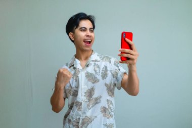 Yazlık kıyafetler giyen mutlu Asyalı genç adam, elinde akıllı telefon tutarken başarıyı kutluyor. Heyecanlı ve gülümsüyor. Terfi için ideal.