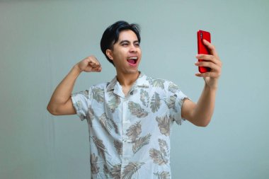 Yazlık kıyafetler giyen mutlu Asyalı genç adam, elinde akıllı telefon tutarken başarıyı kutluyor. Heyecanlı ve gülümsüyor. Terfi için ideal.