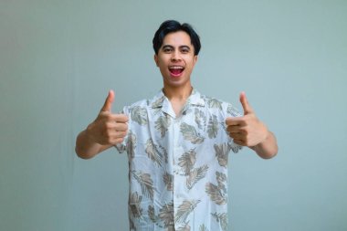 Yazlık giysiler içinde mutlu, genç Asyalı bir adam. Gülümsüyor ve kameraya iki parmağını kaldırıyor. Heyecan ve pozitifliği ifade ediyor.