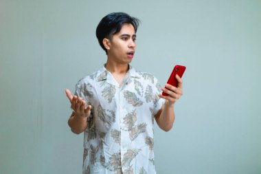 Yazlık kıyafetli genç Asyalı bir adam elinde akıllı bir telefonla, net bir arkaplan karşısında şaşkınlıkla tepki veriyor, dijital reklamlar, mobil uygulamalar veya promosyon içeriği için mükemmel.