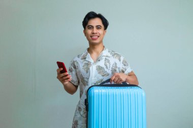 Akıllı telefon kullanırken gülümseyen ve mavi bir bavul taşıyan, seyahat ya da tatile hazır, temiz bir arka planda izole edilmiş, mutlu, Asyalı genç adam.