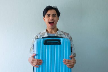 Yazlık giysiler içinde mutlu genç Asyalı bir adam, bavula sarılmış, etkileyici bir ifadeyle, kameraya bakıyor, izole edilmiş, tatile ya da seyahate hazır.