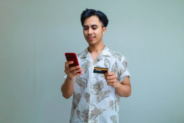 Elinde kredi kartı olan ve cep telefonu kullanan gülümseyen Asyalı genç bir adam rahat ve neşeli bir ruh hali sergiliyor.