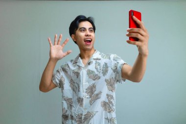 Yazlık kıyafetli mutlu genç Asyalı adam kendini akıllı bir telefonla kaydediyor, gülümsüyor ve temiz bir geçmişe karşı eğlenceli bir jest yapıyor, sosyal medya veya reklam kullanımı için mükemmel.