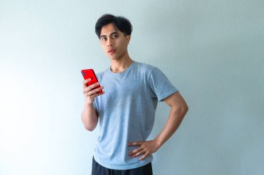 Ciddi bir Asyalı genç adamın portresi. Bir eli belinde duruyor, cep telefonu kullanıyor, temiz bir arkaplanda izole edilmiş, internet reklamları veya iletişim temaları için mükemmel.