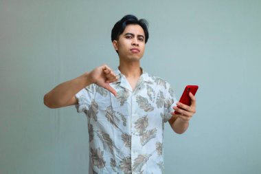 Yazlık kıyafetleri içinde ciddi bir Asyalı adam elinde akıllı telefon ve baş parmağını kaldırma hareketi yapıyor, temiz bir arka plana karşı duruyor, onaylamadığını ya da olumsuz geri bildirim yaptığını söylüyor.