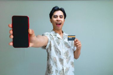 Yazlık giysiler içinde, elinde boş bir telefon ekranı ve temiz bir arka planda izole edilmiş ürün reklamı için bir kart tutan mutlu bir Asyalı.