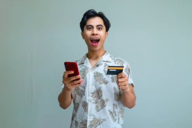 Yazlık kıyafetli mutlu bir Asyalı adam elinde bir kredi kartı tutuyor ve internet üzerinden işlem için akıllı telefon kullanıyor, net bir arkaplanda şaşkın yüz ifadesi gösteriyor.