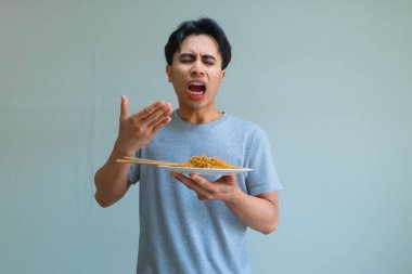 Bir tabak erişte yiyen genç Asyalı adam izole edilmiş arka planda komik bir yüz ifadesi sergiliyor, neşeli ve anlamlı bir yemek tepkisi yakalıyor.