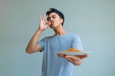 Genç Asyalı bir adam, mutlu ve tatmin edici bir ifadeyle lezzetli eriştelerin tadını çıkarıyor, yemek yerken dudaklarını yalıyor, arka planda izole bir şekilde.