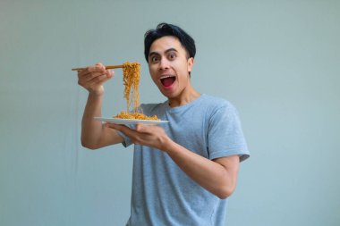 Yemek çubuklarıyla erişte yiyen mutlu genç Asyalı adam temiz bir arka planda heyecanlı bir yüz ifadesi sergiliyor. Yemek, kültür ya da yaşam tarzı teması için mükemmel.