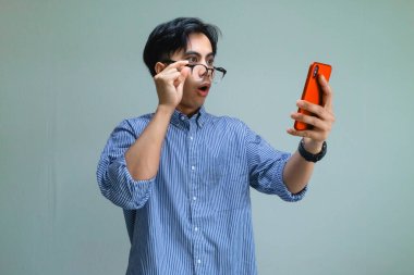 Şok olmuş Asyalı genç bir adam inançsızlıkla akıllı telefon ekranına bakıyor, arka planda izole edilmiş, sürpriz ya da beklenmedik haberler veriyor, dijital, teknoloji ya da reklam kullanımı için mükemmel.