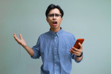 Sinirli Asyalı genç bir adam elinde akıllı bir telefon tutarak hayal kırıklığını ya da öfkesini ciddi bir suratla ifade ediyor. Sade bir zeminde izole edilmiş. Dijital sorunlar veya reklam kavramları için mükemmel