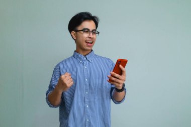 Akıllı telefonu tutarak başarıyı kutlayan mutlu Asyalı genç adam, izole edilmiş arka planda heyecanlı bir şekilde gülümsüyor, dijital reklamlar veya promosyonlar için mükemmel.