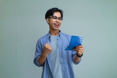 Mutlu Asyalı genç adam zafer jesti ile başarıyı kutlarken bir tasarruf kitabına bakıyor. Sade bir arka plan, finansal ve başarı kavramına izole edilmiş.