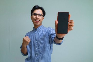 Mutlu Asyalı genç adam, boş bir akıllı telefon ekranı göstererek başarıyı kutluyor, izole bir arkaplanda zafer jesti yapıyor, reklam ve promosyonlar için mükemmel.