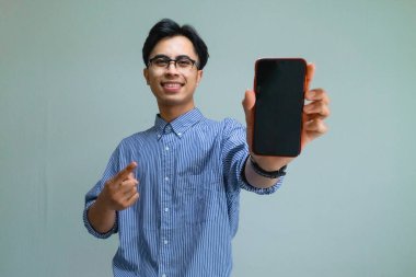 Neşeli Asyalı genç adam elinde boş ekran ile akıllı telefonu tutuyor, arka planda kameraya gülümsüyor, teknoloji, uygulama veya reklam kavramları için mükemmel.