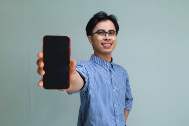 Neşeli Asyalı genç adam elinde boş ekran ile akıllı telefonu tutuyor, arka planda kameraya gülümsüyor, teknoloji, uygulama veya reklam kavramları için mükemmel.
