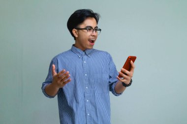 Zeki telefonuna şaşırmış bir ifadeyle bakan mutlu genç Asyalı adam, arka planda izole edilmiş, ekrandaki bir şeye tepki verirken heyecan ve neşe gösteriyor.