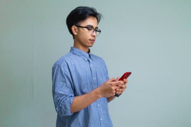 Ciddi Asyalı genç adam yazarken akıllı telefonuna odaklanmış, temiz bir arkaplanda izole edilmiş.