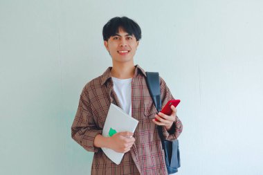 Sırt çantası taşıyan ve elinde cep telefonu tutan bir üniversite öğrencisi kameraya bakarken gülümsüyor.