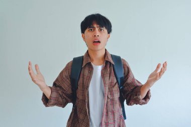 Şok olmuş erkek öğrenci sırt çantası takıyor. Kafası karışmış bir şekilde kameraya bakıyor. İki elini de kaldırıyor.