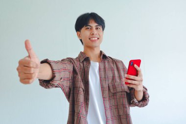 Genç Asyalı adam gülümsüyor ve beyaz bir arka planda izole edilmiş bir cep telefonunu tutarken başparmağını kaldırıyor.