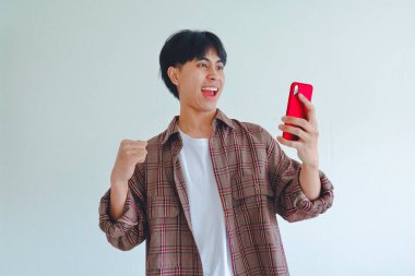 Heyecanlı Asyalı adam telefona bakarken elini kaldırıyor, başarıyı neşeli bir ifadeyle kutluyor, sade bir arka planda izole edilmiş..