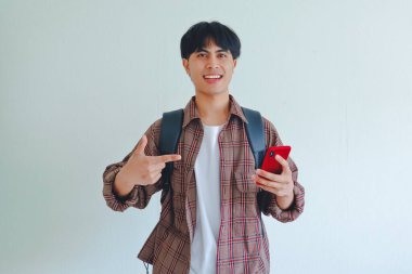Sırt çantalı gülümseyen Asyalı erkek öğrenci telefonu tutuyor ve işaret ediyor, beyaz arka planda izole edilmiş, eğitim veya teknoloji reklamları için mükemmel.