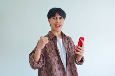 Heyecanlı Asyalı adam telefona bakarken elini kaldırıyor, başarıyı neşeli bir ifadeyle kutluyor, sade bir arka planda izole edilmiş..