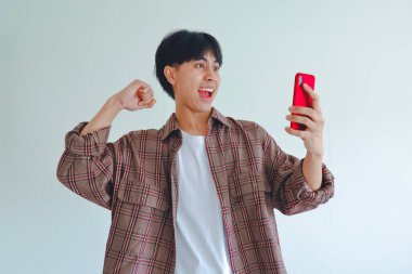 Heyecanlı Asyalı adam telefona bakarken elini kaldırıyor, başarıyı neşeli bir ifadeyle kutluyor, sade bir arka planda izole edilmiş..