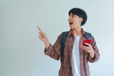 Sırt çantası taşıyan bir erkek üniversite öğrencisi yanındaki boşluğu işaret ediyor ve cep telefonunu tutarken şaşırmış bir ifade sergiliyor..