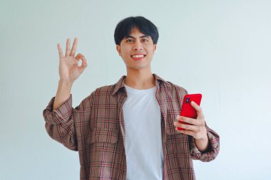 Mutlu Asyalı adam akıllı telefon tutarken tamam işareti gösteriyor ve beyaz arka planda gülümsüyor, reklam için izole edilmiş stüdyo portresi